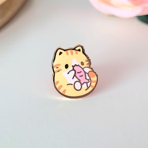 May include: A gold-colored enamel pin featuring a cartoon cat holding a pink fish. The cat has orange stripes and a white belly. The pin is on a light blue surface, with a wooden tray and a pink rose in the background.