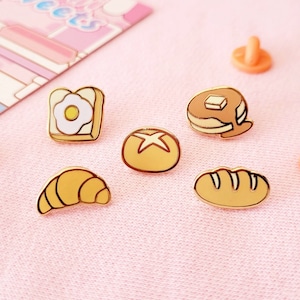 May include: Six gold-coloured enamel pins featuring different breakfast foods: a croissant, a slice of bread with a fried egg, a bun with a cross on top, a loaf of bread, and two pancakes with butter and syrup. The pins are on a pink surface.