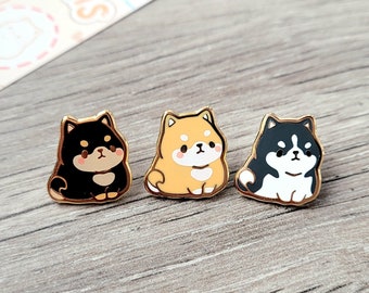 May include: Three enamel pins featuring cartoon Shiba Inu dogs. The dogs are in black, orange, and black and white. The pins are on a woodgrain surface.