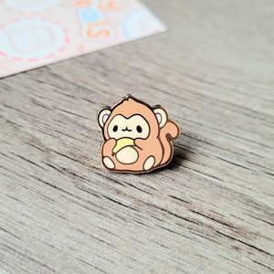 May include: A gold-colored enamel pin featuring a cartoon monkey holding a banana. The monkey is brown with a light pink face and has a small, white tail.