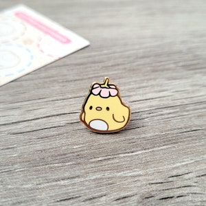 May include: A gold-colored enamel pin featuring a cartoon chick wearing a pink flower on its head. The chick is yellow with white eyes and a white belly.