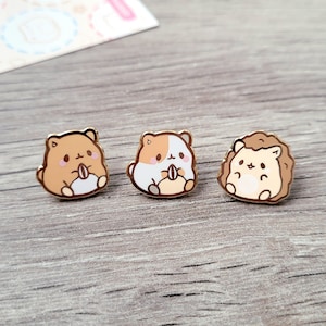 May include: Three enamel pins featuring cartoon animals. The pins are gold-toned and feature a brown hamster, a white hamster, and a brown hedgehog. Each animal is holding a small object in its paws.