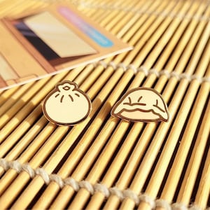 May include: Two gold-colored enamel pins shaped like dumplings. One dumpling is round and the other is half-moon shaped.