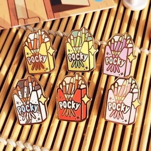May include: A selection of Pocky enamel pins in shades of brown, yellow, green, pink, red, and white. Each pin depicts a Pocky box with the word "Pocky" and a starburst design. The pins are displayed on a bamboo mat.