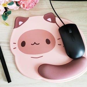 May include: A pink cat-shaped mousepad with a black computer mouse and a black stylus pen. The mousepad has a brown wrist rest and a cartoon cat face design. The mouse is connected to a black cable.