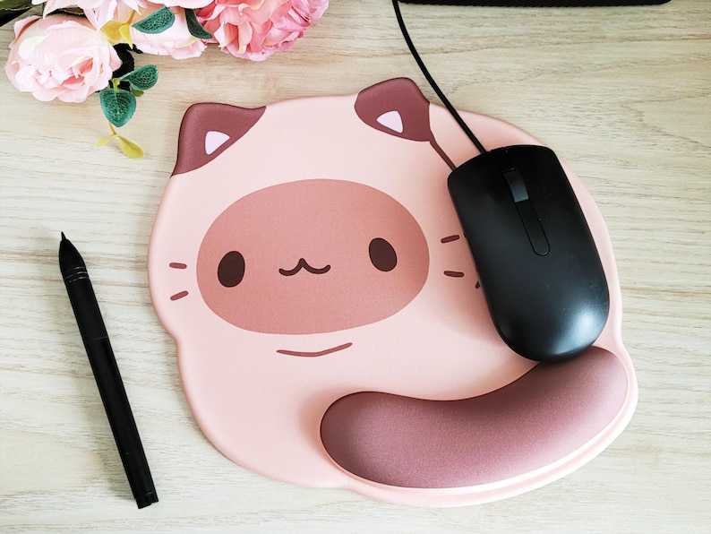 May include: A pink cat-shaped mousepad with a black computer mouse and a black stylus pen. The mousepad has a brown wrist rest and a cartoon cat face design. The mouse is connected to a black cable.