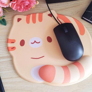 May include: A beige cat-shaped mousepad with orange stripes and accents. A black computer mouse rests on the pad. A black stylus and a notebook are also visible. Pink flowers are in the background.
