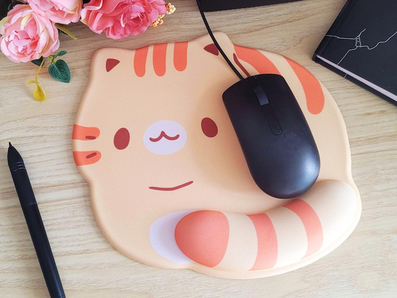 May include: A beige cat-shaped mousepad with orange stripes and accents. A black computer mouse rests on the pad. A black stylus and a notebook are also visible. Pink flowers are in the background.