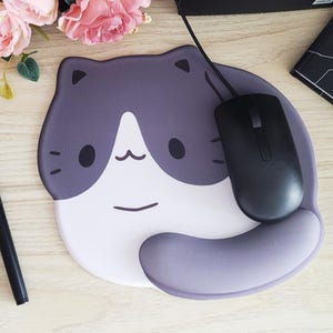 May include: A cat-shaped mousepad with a black mouse. The mousepad is gray and white with a cartoon cat face design. A black pen and pink flowers are also on the wooden surface.