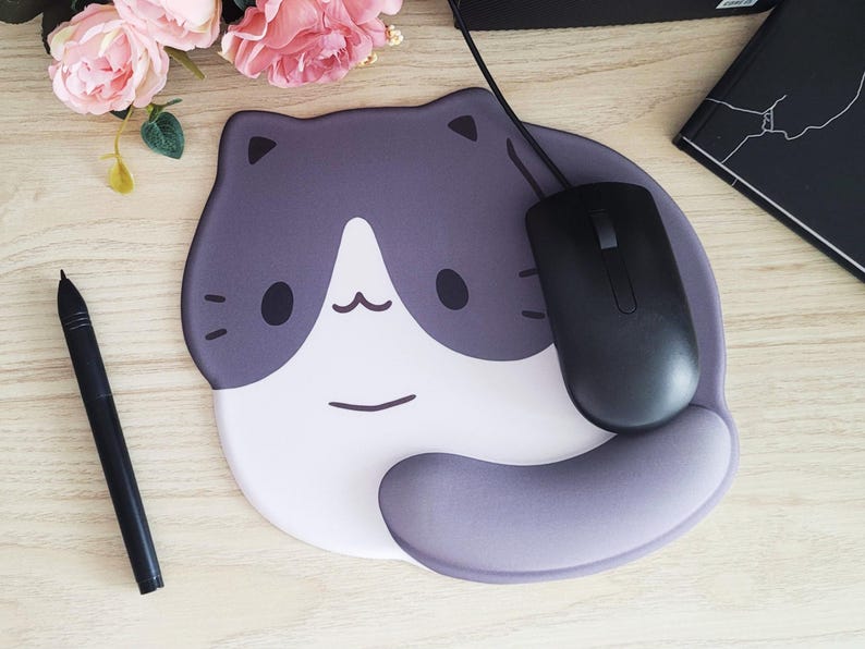 May include: A cat-shaped mousepad with a black mouse. The mousepad is gray and white with a cartoon cat face design. A black pen and pink flowers are also on the wooden surface.