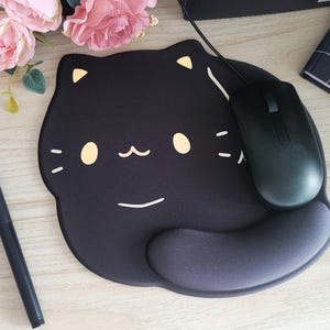 May include: Black cat-shaped mousepad with a black mouse. The mousepad has a wrist rest and a cute cat face design with yellow accents. A black pen and pink flowers are also in the image.