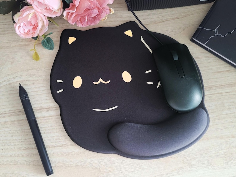 May include: Black cat-shaped mousepad with a black mouse. The mousepad has a wrist rest and a cute cat face design with yellow accents. A black pen and pink flowers are also in the image.
