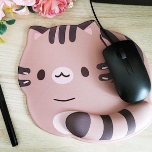 May include: A pink cat-shaped mousepad with a black computer mouse. The mousepad has a cartoon cat face design with brown stripes and details. A black pen is also visible on the wooden surface.