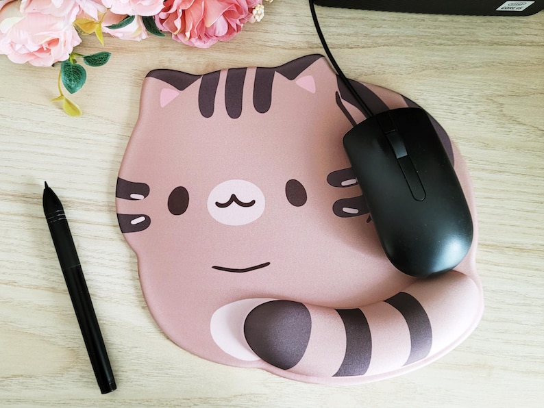 May include: A pink cat-shaped mousepad with a black computer mouse. The mousepad has a cartoon cat face design with brown stripes and details. A black pen is also visible on the wooden surface.