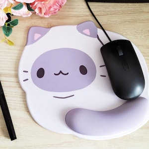 May include: A white and lavender cat-shaped mousepad with a black computer mouse and a black stylus pen. The mousepad has a lavender face and ears, with a lavender wrist rest. Pink flowers are in the background.