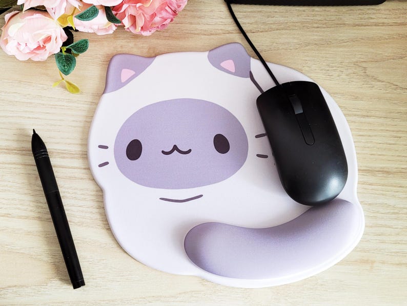 May include: A white and lavender cat-shaped mousepad with a black computer mouse and a black stylus pen. The mousepad has a lavender face and ears, with a lavender wrist rest. Pink flowers are in the background.