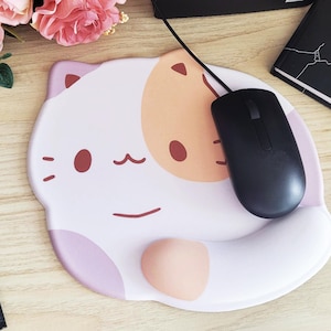 May include: A white and pink cat-shaped mousepad with a black mouse. The mousepad has a wrist rest and a cartoon cat face design with brown eyes and a smiling mouth. A black pen and a notebook are also visible.