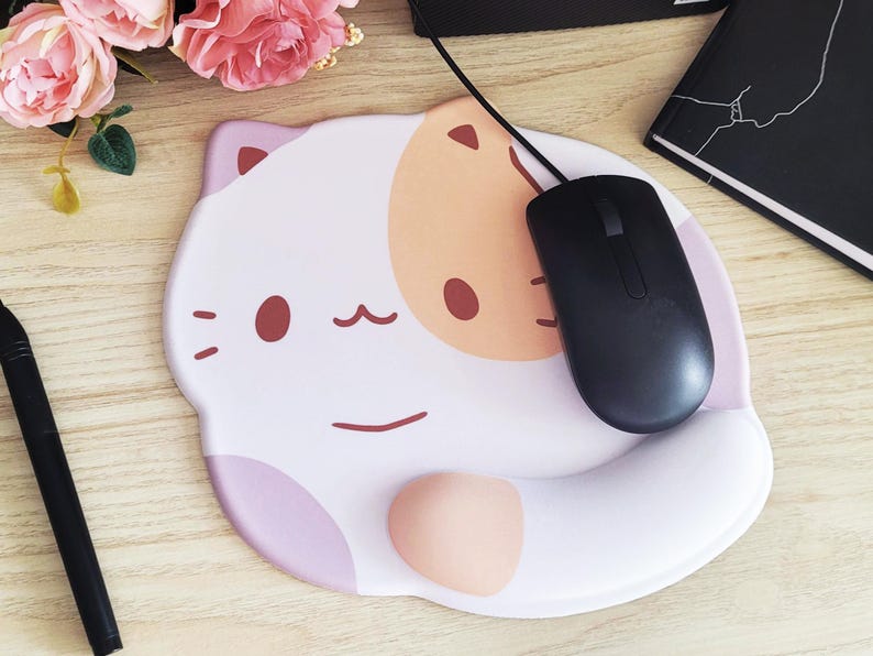 May include: A white and pink cat-shaped mousepad with a black mouse. The mousepad has a wrist rest and a cartoon cat face design with brown eyes and a smiling mouth. A black pen and a notebook are also visible.