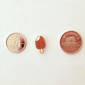 May include: A gold-colored ice cream pin with a brown chocolate coating and white ice cream. The pin is next to two Canadian coins, one is a 10-cent coin from 2014 and the other is a 5-cent coin from 2018.