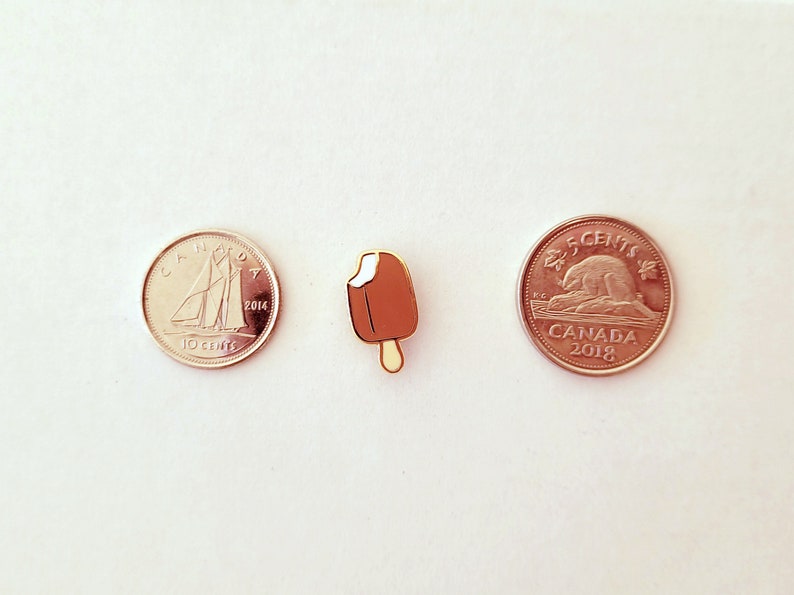 May include: A gold-colored ice cream pin with a brown chocolate coating and white ice cream. The pin is next to two Canadian coins, one is a 10-cent coin from 2014 and the other is a 5-cent coin from 2018.