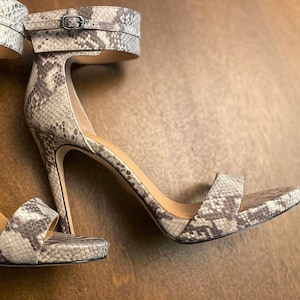 Peut inclure: Sandales à talons hauts avec un motif peau de serpent gris et marron. Les sandales ont une fine bride de cheville avec une boucle et une seule bride sur les orteils. Les talons sont fins.