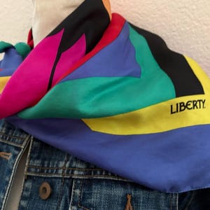 May include: A multi-colored silk scarf featuring bold geometric sections in magenta, green, blue, yellow, red, and black. The word LIBERTY is printed in black capital letters on a yellow section. The scarf is draped over a blue denim jacket.