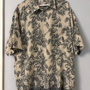 Pode incluir: Uma camisa de manga curta com botões em tom bege claro, apresentando um padrão de folhas tropicais em cinza carvão escuro. A camisa inclui uma gola pontiaguda, um bolso no peito e uma faixa decorativa horizontal perto da bainha inferior.