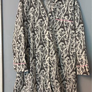 May include: A white button-down nightshirt featuring a repeating dark gray pattern of the Eiffel Tower and floral sprigs. The garment includes a collared neckline, long sleeves with pink piping on the cuffs, and a single chest pocket with pink trim.