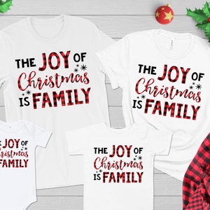 May include: White t-shirts with the text "THE JOY OF Christmas IS FAMILY" in black and red plaid lettering. The shirts are in various sizes, including a baby onesie. A red and black plaid shirt is visible.