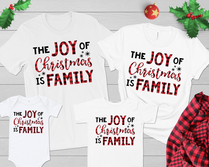 May include: White t-shirts with the text "THE JOY OF Christmas IS FAMILY" in black and red plaid lettering. The shirts are in various sizes, including a baby onesie. A red and black plaid shirt is visible.