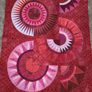 May include: A handmade quilt in shades of red, pink, and burgundy. The quilt features a geometric design with circular and semi-circular shapes, each with a spiky, sunburst-like pattern. The background is a solid red with a subtle polka dot texture.