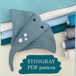 May include: A handmade felt stingray in shades of gray, with white spots and black eyes. The image includes spools of thread and stacks of felt in various shades of blue and teal. Text on a teal banner reads "STINGRAY PDF pattern."