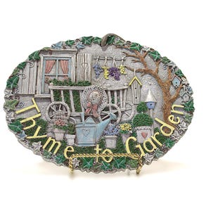May include: Oval garden-themed plaque with the words "Thyme to Garden" in gold lettering. The plaque features a miniature garden scene with a cart, watering can, and potted plants. The border is decorated with green ivy.