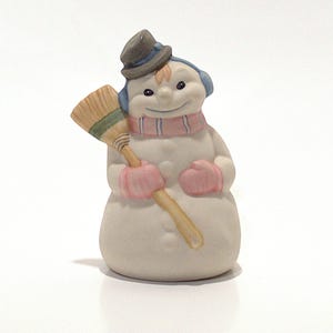 May include: A white ceramic snowman figurine with a blue hat, pink mittens, and a striped scarf holding a brown broom.