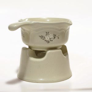 May include: A beige ceramic fondue pot with a single handle and a matching base stand. The pot features a subtle speckled texture and a small, grey floral pattern printed on the side. The base has a wide, circular opening at the top.