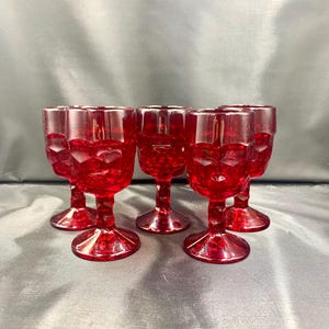 Könnte beinhalten: Set aus fünf rubinroten Vintage-Likörgläsern. Jedes Glas hat ein strukturiertes, geometrisches Muster auf der Schale und einen zarten Stiel. Die Gläser sind auf einer dunklen Oberfläche angeordnet, um ihre lebendige Farbe und ihr Design hervorzuheben.