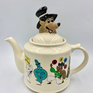 Puede incluir: Una tetera de cerámica blanca con un perro de dibujos animados que lleva un sombrero de copa negro como tapa. La tetera tiene un diseño de dibujos animados colorido con un perro haciendo malabares con pelotas, una botella y un fantasma que sostiene un cartel que dice "Me encanta Jim Beam".