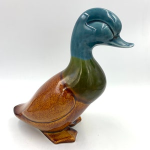 Puede incluir: Una figurita de pato de cerámica con un esmalte marrón, verde y azul. El pato está sentado con la cabeza girada hacia un lado.