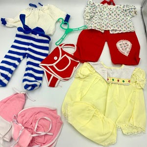 Könnte beinhalten: Eine Sammlung von Vintage-Puppenkleidung in verschiedenen Stilen und Farben. Enthält einen weißen Pullover mit blauen Verzierungen, blau-weiß gestreifte Hosen, rote Hosen, ein Oberteil mit Blumenmuster, eine rote Handtasche und gelbe Kleider. Enthält auch rosa und weiße Puppenaccessoires.