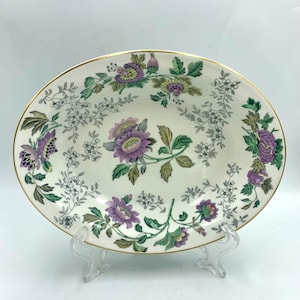 Könnte beinhalten: Eine ovale Servierschale aus Keramik mit floralem Design. Die Schale hat einen cremefarbenen Grund mit lila und grünen Blumenmustern und einen goldenen Rand. Die Schale wird auf einem transparenten Kunststoffständer präsentiert.