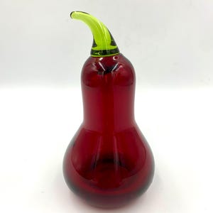 Puede incluir: Una pieza de arte decorativo de vidrio con forma de pimiento. El cuerpo es de color rojo intenso, con un tallo curvo de color verde brillante. El vidrio tiene un acabado brillante y se asienta sobre una superficie blanca.