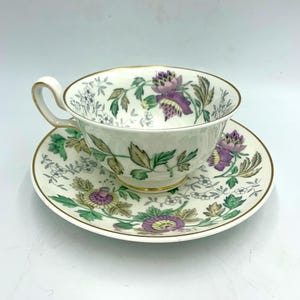 May include: A white teacup and saucer set with a floral design. The teacup and saucer have a gold trim. The floral design includes purple, green, and brown leaves and flowers. The teacup has a curved handle.