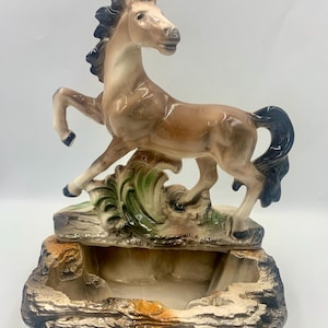 May include: A ceramic horse figurine in shades of brown, tan, and black, rearing up on a base with a trough. The horse has a flowing mane and tail, with green and white accents on the base. The base has a rock-like texture.