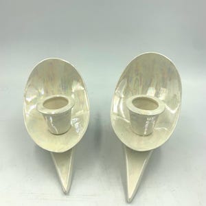 Pode incluir: Um par de castiçais de cerâmica branca com um design único e abstrato. Cada castiçal apresenta uma base curva de forma oval com uma ponta pontiaguda e uma pequena taça redonda para velas.