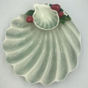 May include: A ceramic serving dish shaped like a seashell with a smaller shell-shaped dip in the center. The dish is decorated with green leaves and red strawberries.