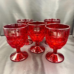 Puede incluir: Conjunto de cinco copas de vino tinto rubí. Cada copa presenta un diseño facetado en el cuenco y una base redonda. Las copas son de material transparente y están dispuestas sobre una superficie oscura.