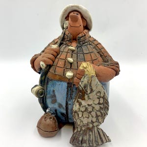 May include: A ceramic figurine of a fisherman wearing a plaid shirt, blue pants, and a white hat. The fisherman is holding a fishing rod and a fish. The figurine is brown, blue, and white.