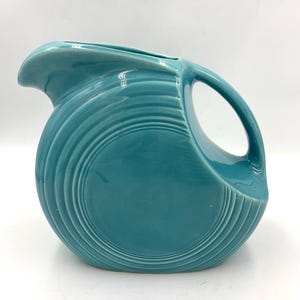May include: A turquoise ceramic pitcher with a distinctive circular design and a large handle. The pitcher has a spout for pouring and is likely used for serving beverages. The surface features concentric, ridged detailing.
