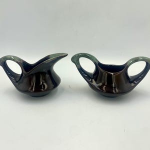 May include: Two ceramic serving dishes with a dark brown glaze and green handles. One dish has a spout, and the other has two handles. The dishes are on a white background.