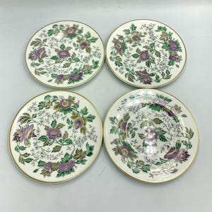 May include: Four white ceramic plates with a floral design featuring purple and green flowers and foliage. The plates have a gold rim and are arranged in a square.